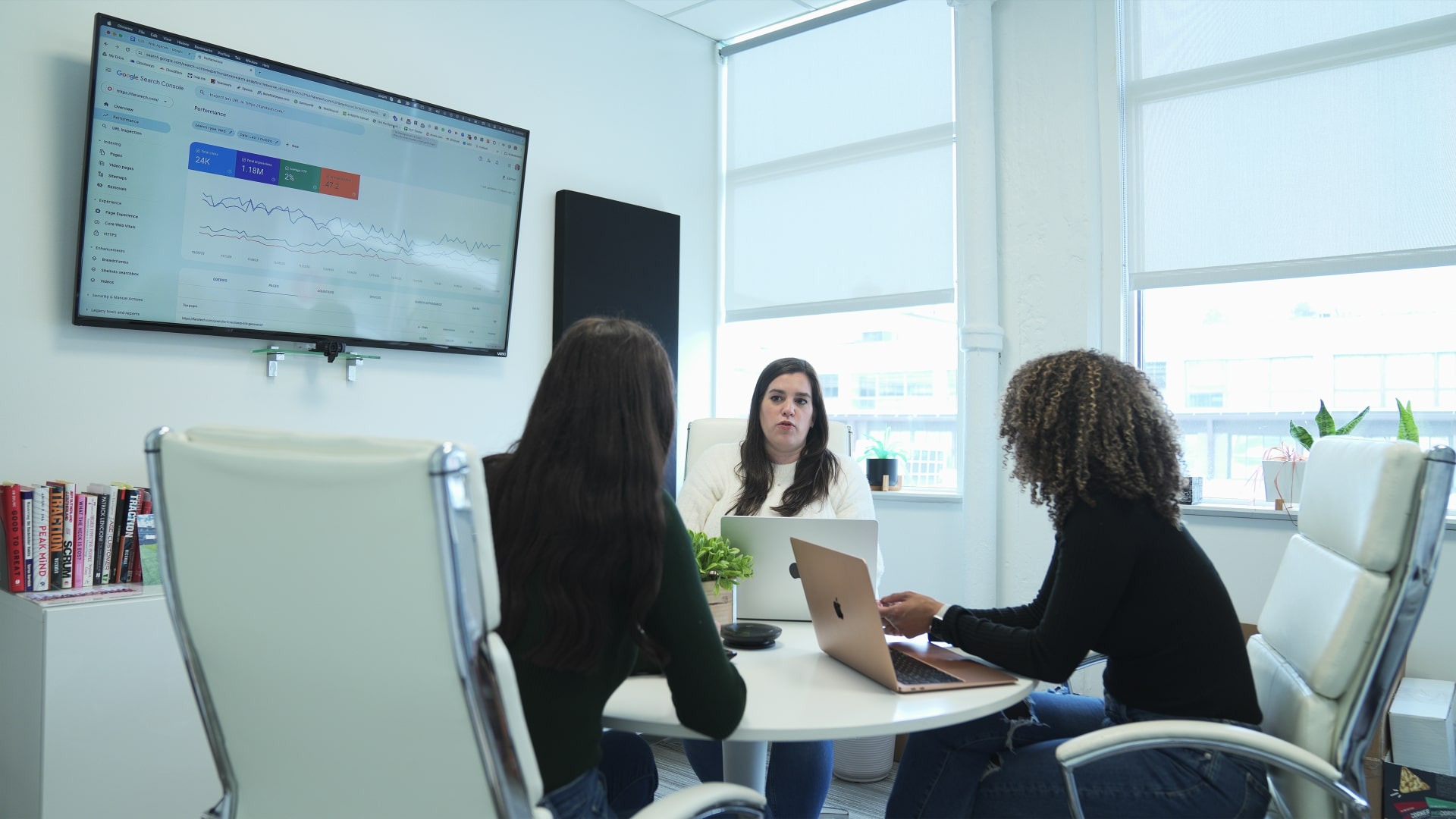 Three women sit around a small round table with open laptops while they discuss marketing analytics, which are displayed on a screen on the wall